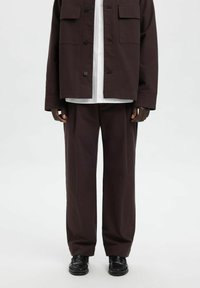 Mørkebrun jakke med løs passform og to brystlommer med svarte knapper, kombinert med matchende vide bukser og svarte sko.