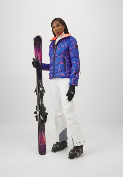 Blauer Puffermantel mit pinkem Rand, weiße Skihosen, schwarze Handschuhe und schwarze Skischuhe. Hält einen lila Ski mit einer schwarzen Bindung.