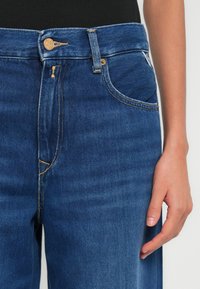 Σκούρο μπλε τζιν με ψηλή μέση, κλείσιμο με κουμπιά και αντίθετη κίτρινη ραφή. Διαθέτει τσέπες και λεία υφή.