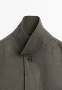 Nahaufnahme einer dunkelgrauen Jacke mit hohem Kragen, die mit einem einzelnen schwarzen Knopf geschlossen wird und sichtbare Nahtdetails an den Schultern aufweist.