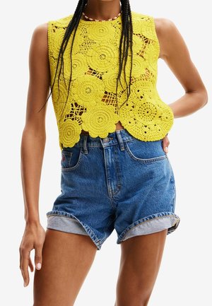Gelbes gehäkeltes Crop-Top mit Blumenmustern, kombiniert mit blauen Denim-Shorts mit umgeschlagenen Säumen und einer Fronttasche.