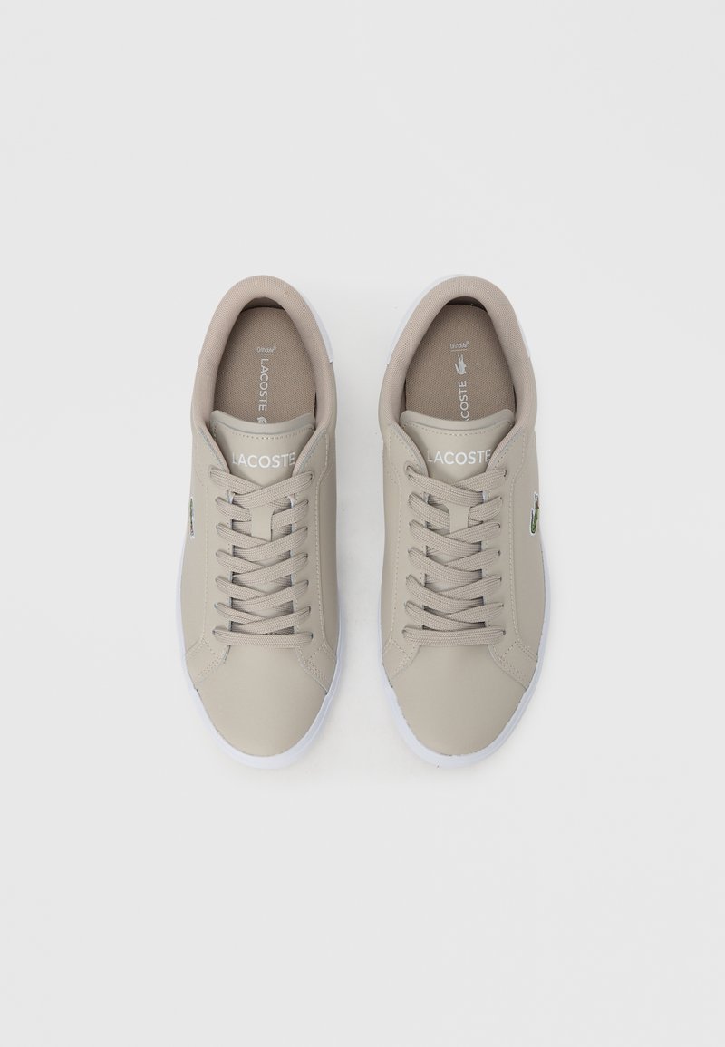 Lys beige lædersneakers med en glat tekstur, rund tå, hvid gummisål og grå snørebånd. Lacoste-logo vist på siden.