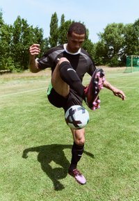 Black athletic shirt with green accents, black shorts, and black socks. Player juggling a soccer ball on grass, wearing colorful cleats.