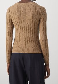 Lichtbruine kabelgebreide trui met lange mouwen, ribbelzoom en textuurpatroon. Draag met donkerblauwe broek, deze is van achteraf gezien.