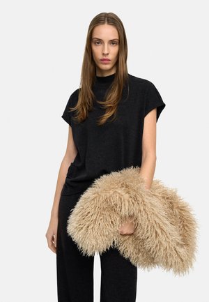 Kobieta z długimi brązowymi włosami, ubrana w czarną bluzkę z krótkim rękawem i spodnie, trzymająca dużą beżową, puszystą poduszkę na białym tle.