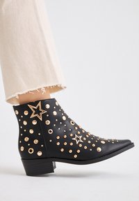Botín negro de cuero, con tachuelas doradas y acentos en forma de estrella, punta afilada y un bajo tacón tipo bloque. Detalle de superficie texturizada.