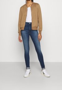 Veste bomber matelassée beige à col rond, assortie à un jean skinny bleu et des baskets blanches avec des accents noirs.
