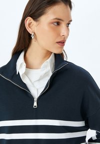 Maglione navy con colletto alto e zip quarto, caratterizzato da strisce orizzontali bianche sul petto e colletto a costine.