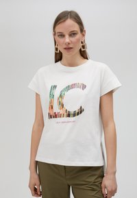 Camiseta blanca de algodón de manga corta, con un logo "LC" multicolor formado por rayas verticales y texto debajo. Presenta aberturas laterales.