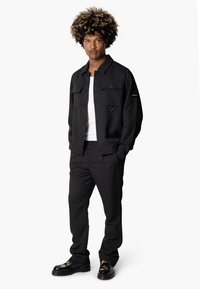 Zwarte jas met twee voorzakken en knopen, gecombineerd met bijpassende broek en zwarte schoenen met een gemusterde print.