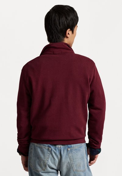 Bordeaux fleece jas met een hoge kraag, zichtbare stiksels en geribde manchetten, gecombineerd met lichtblauwe denim jeans. Achteraanzicht.