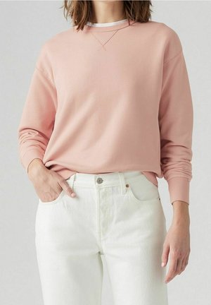 Personne portant un sweat-shirt à manches longues rose clair et un jean blanc taille haute, une main dans la poche sur un fond uni.