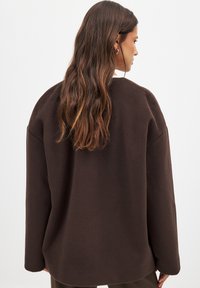 Pruun fliisist sviiter, millel on lahtine lõige, langevad õlad ja pikad varrukad. Tekstuur on pehme ja sile, ümar kaelus.