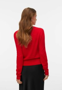 Sarkans adīts džemperis ar garām piedurknēm, ribotām malām un apakšmalu, valkāts virs melnas svārku. Tam ir smalka tekstūra un pieguļošs dizains.