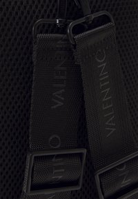 Juodas tinklinis audinys su tekstūruotu diržu, kuriame įspaustas "Valentino". Apima patvarius plastiko lydinius ir reguliuojamus juodus segtukus.