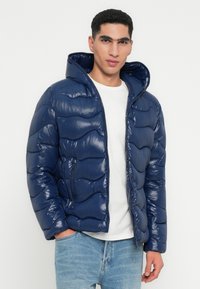 Jachetă puffer bleumarin cu un finisaj lucios, dotată cu glugă, model de cusături în zig-zag și buzunare cu fermoar pe laterale, purtată peste un maiou alb.