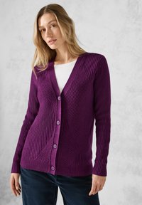 Fialový žebrovaný cardigan s výstřihem do V, který má strukturované vzory a ladící knoflíky, kombinovaný s bílým topem a tmavými kalhotami.
