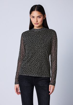 Junge Frau mit langen, dunklen Haaren, die ein schwarzes, durchsichtiges Langarmoberteil mit einem kleinen weißen Blumenmuster und dunklen Jeans trägt, steht vor einem einfarbigen Hintergrund.