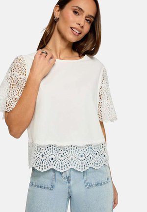 Femme souriante portant un haut blanc avec manches et bas en dentelle, associé à un jean en denim bleu clair, posant devant un fond uni.