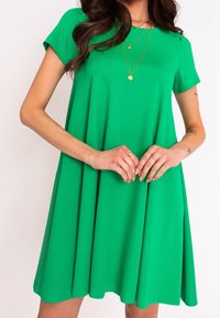 Grünes Kurzarmkleid aus glattem Stoff, mit ausgestellter Form und knielangem Saum. Betont mit geschichteten goldenen Halsketten.