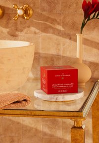 Scatola quadrata rossa etichettata "ricarica di crema corpo soffice" su una piattaforma in pietra marmorizzata. Presenta accessori dorati e un asciugamano beige texture nelle vicinanze.