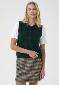 Grüne Strickweste mit Knopfverschluss, über einem weißen kurzärmeligen Hemd getragen, kombiniert mit einem karierten braunen Rock. Strukturiertes und geschichtetes Design.