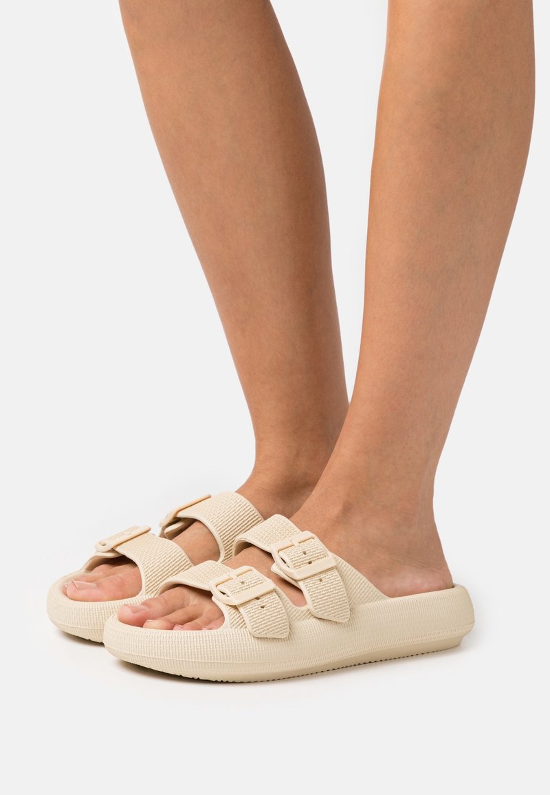Pieds portant des sandales beige à enfiler avec deux brides réglables à boucle sur fond blanc.