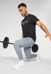 Sort Nike t-shirt med hvid "Just Do It" logo, grå træningsbukser og hvide træningssko. En vægtstang er placeret i nærheden.