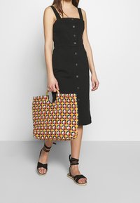 Kobieta w czarnej sukience zapinanej na guziki i czarnych sandałach związywanych na kostce, trzymająca wielokolorową, plecioną torbę na ramię z okrągłymi drewnianymi uchwytami.