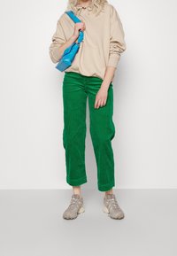 Pantalons en velours côtelé verts, hoodie oversized beige et baskets grises. Un sac bleu pend à l'épaule. Design simple avec un tissu texturé.