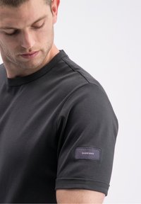 Juoda trumpomis rankovėmis marškinėliai iš lygaus audinio, su apvalia iškirpte ir tekstūruotu ženkliuko lopu kairėje rankovėje.