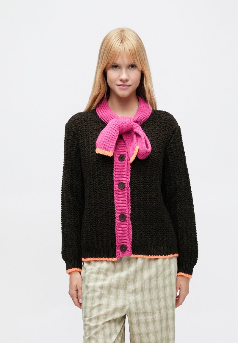 Pletený kardigán tmavohnedej farby s jasne ružovým rebrovaným predným dielom, ozdobený oranžovým lemom a gombíkmi. Nosený cez kárované nohavice.