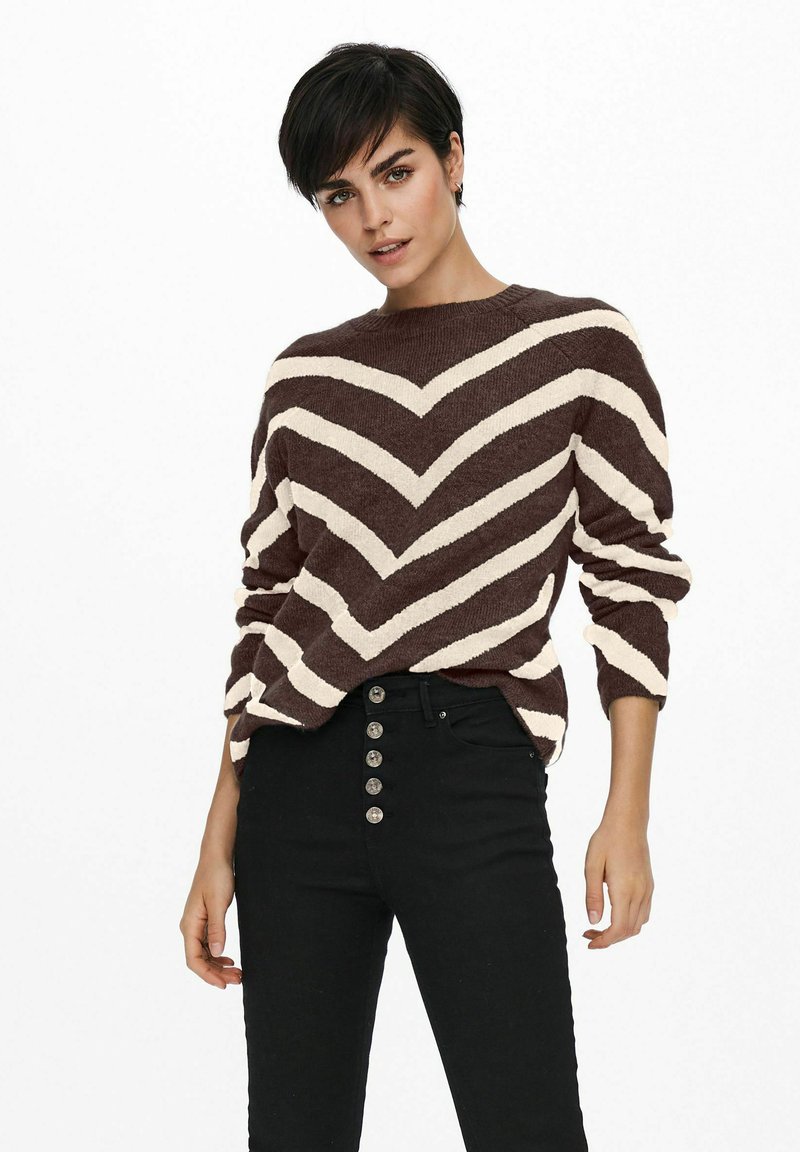 Femme aux cheveux courts et foncés, portant un pull à rayures en chevron marron et blanc ainsi que des pantalons noirs taille haute avec des boutons.