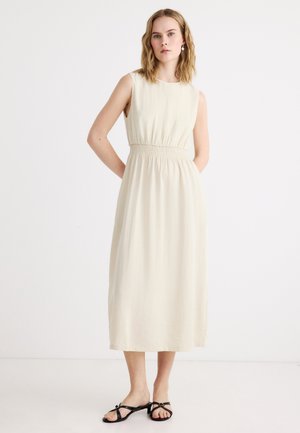 Vrouw met licht golvend haar, gekleed in een mouwloze offwhite midi-jurk met een geplooide taille en zwarte sandaal met hakken, staand tegen een witte achtergrond.