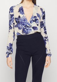 Blusa floreale fluida color crema e blu con maniche lunghe e scollo a V profondo; abbinata a pantaloni aderenti a coste scuri con cuciture curve.