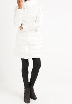 Femme portant un long manteau matelassé blanc avec une fermeture éclair, un pantalon slim noir et des bottines noires, debout avec une main près du visage.