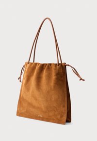 Sac fourre-tout en suède marron clair avec fines bretelles en cuir et détails à cordon latéral, présenté sur un fond blanc.