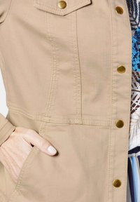 Beige jas gemaakt van gladde stof, met een slim fit, knoopsluitingen in messing en zijzakken. Zichtbare stiksels accentueren de naden.