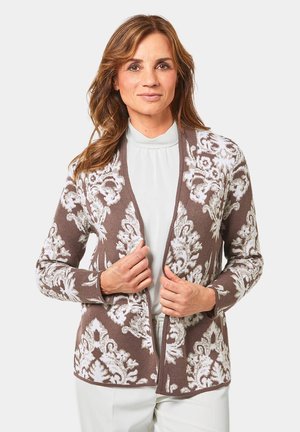Bruine cardigan met witte bloempatronen, open voorkant, lange mouwen en een zachte textuur, gecombineerd met een witte top.