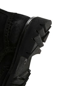 Bota negra de ante con una suela de goma texturizada, que presenta un patrón en zigzag y acentos perforados a lo largo del borde superior.