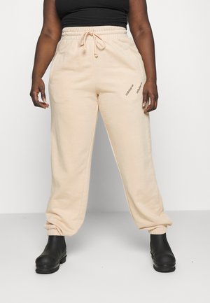 Osoba nosząca beżowe spodnie dresowe Adidas z regulowanym sznurkiem w talii oraz czarne botki, stojąca na tle jednolitego białego tła.