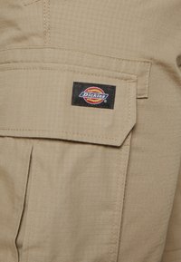 Poche cargo en tissu beige avec rabat et petite étiquette noire rectangulaire ornée du logo coloré Dickies cousue dessus.