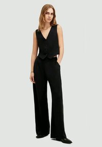 Colete preto ajustado com decote em V e fecho de botão, combinado com calças pretas de perna larga de cintura alta. Textura de tecido suave.