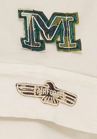 Vit tyg med en broderad grön och blå "M" logotyp samt en brun "AIRFORCE" text tillsammans med en örn-emblem. Detaljerad sömnad är synlig.