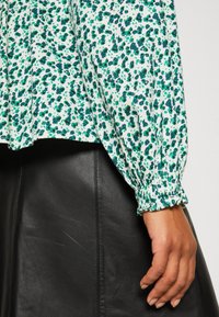 Bluse mit floralen Mustern in Grün und Marine auf weißem Hintergrund, ausgestattet mit langen, gerafften Ärmeln und lockerer Passform, kombiniert mit einem schwarzen Lederrock.