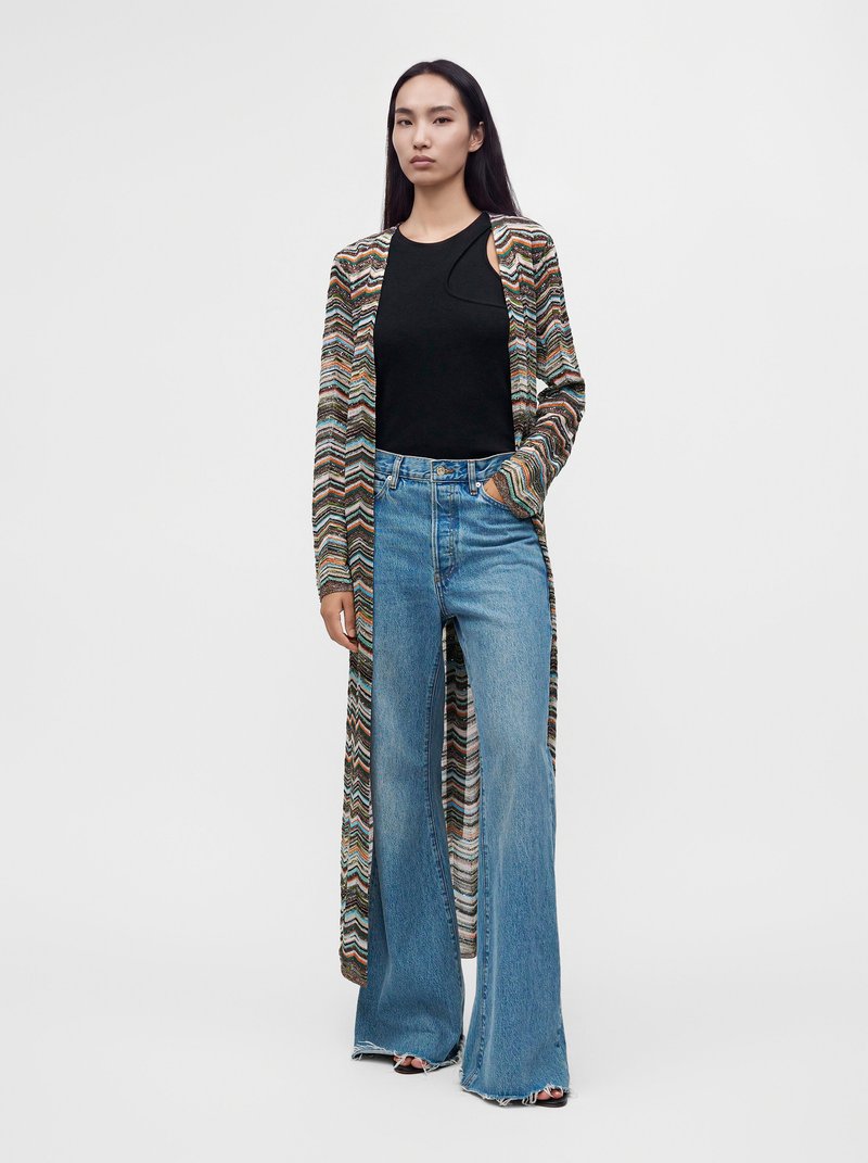 Femme portant un long cardigan à motifs, un top noir sans manches et un jean bleu taille haute à jambes larges sur un fond uni.