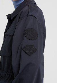 Chaqueta azul marino con un diseño estructurado, que presenta dos parches negros bordados en la manga y un bolsillo delantero abotonado.