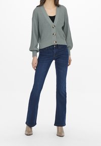 Cardigan en tricot vert clair avec un décolleté en V profond, trois grands boutons et des manches ballons, associé à un jean évasé bleu foncé et des bottes beiges.