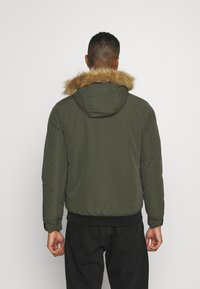 Giacca puffer in verde oliva con cappuccio foderato in pelliccia sintentica, polsini e orlo a coste, caratterizzata da una texture liscia e un design minimalista.