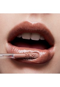Gloss à lèvres liquide avec une couleur marron nude brillant appliqué sur des lèvres pleines ; un applicateur transparent est utilisé pour l'application. Texture lisse visible.